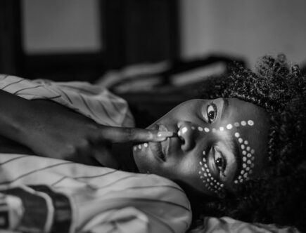 Selective Focus Grayscale Photo of Women in Face Paint Lying Down Posing With Her Finger on Her Lips
