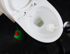 From above crop anonymous person in yellow gloves cleaning toilet bowl with toilet brush and liquid detergent in green plastic bottle